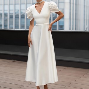 A white midi dress with a V-neckline and short, puffed sleeves. The dress features a fitted waist and a flared skirt. The person is wearing white heels and a pearl choker. The background includes a modern building and a wooden deck.