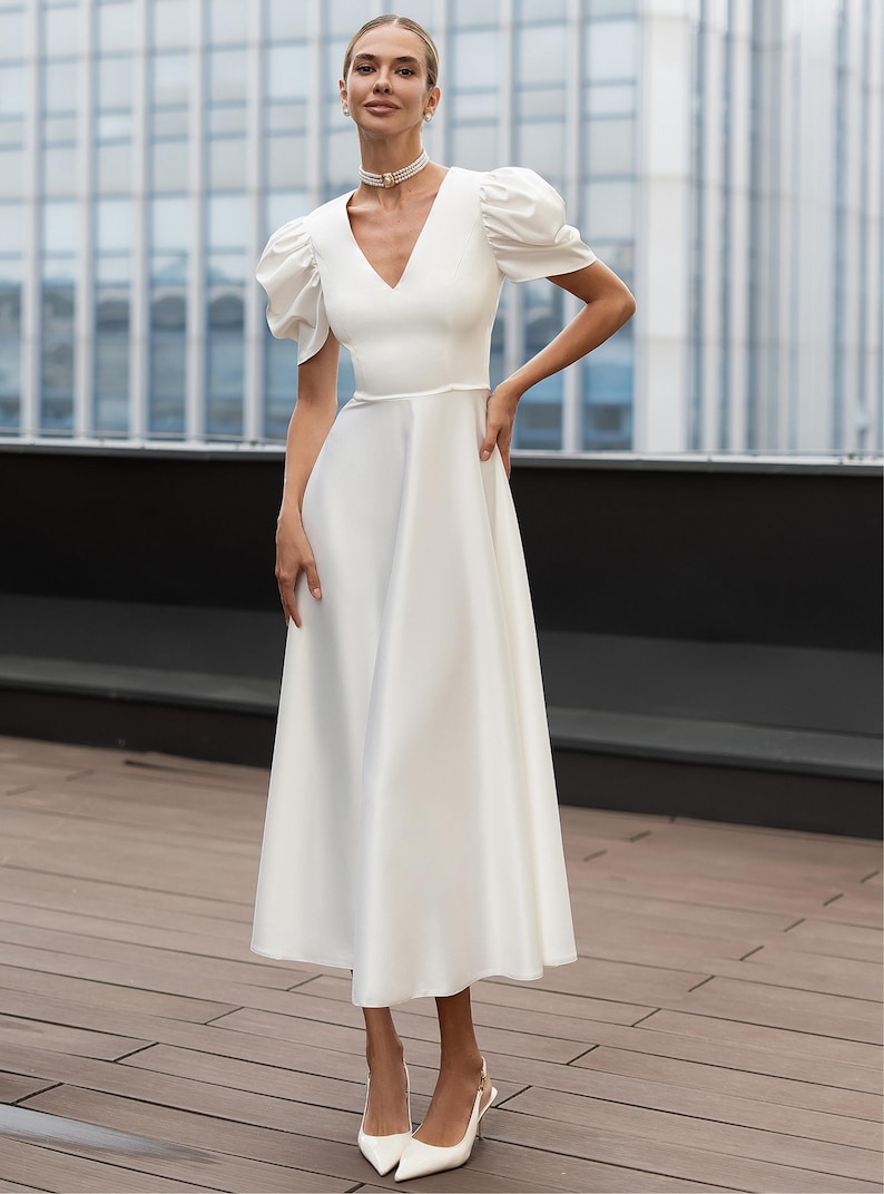 A white midi dress with a V-neckline and short, puffed sleeves. The dress features a fitted waist and a flared skirt. The person is wearing white heels and a pearl choker. The background includes a modern building and a wooden deck.
