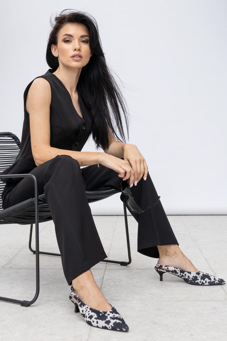 May include: A black sleeveless jumpsuit with a button-down vest and flared trousers. The outfit is paired with black and white patterned kitten heel shoes. The person is seated on a black chair, holding sunglasses.