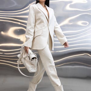 Könnte beinhalten: Cremefarbener Anzug, bestehend aus einem Blazer mit fallendem Revers und passender Hose. Das Outfit wird durch eine Handtasche und Stiefeletten mit Absatz ergänzt. Der Hintergrund ist eine reflektierende, metallische Oberfläche.