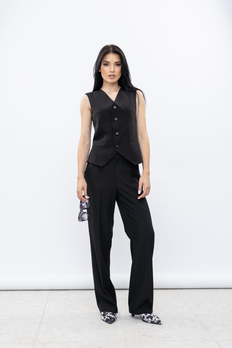 May include: A black waistcoat with a V-neck and button fastening, worn with matching black trousers. The outfit is finished with patterned shoes and sunglasses. The ensemble is a classic, tailored look.