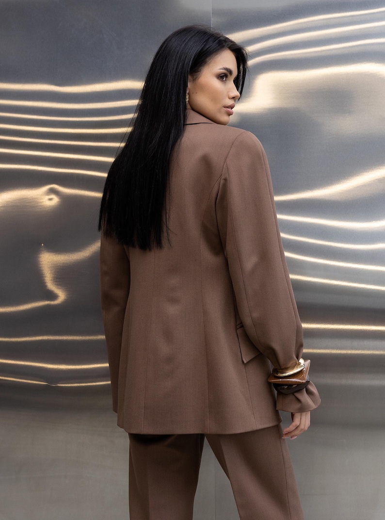 Pu&ograve; includere: Un completo marrone, composto da un blazer e pantaloni, &egrave; mostrato. Il blazer ha un design classico con revers a lancia e maniche lunghe. Le maniche sono raccolte al polso. Il completo &egrave; realizzato in un tessuto liscio. &Egrave; visibile un braccialetto dorato.