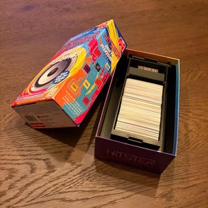 May include: A HITSTER game box, open to show a deck of cards. The box is brightly coloured with red, blue, and yellow. The game title and the words "GUILTY PLEASURE" are visible. The game is on a wooden surface.