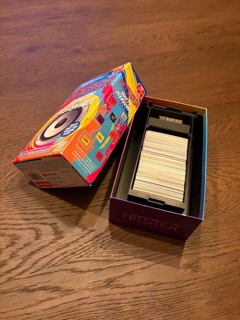 May include: A HITSTER game box, open to show a deck of cards. The box is brightly coloured with red, blue, and yellow. The game title and the words "GUILTY PLEASURE" are visible. The game is on a wooden surface.