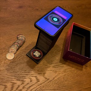 May include: A smartphone showing a colourful circular graphic is propped on a black stand. A stack of circular game pieces with the word "HITSTER" are next to the stand. An open box with the word "HITSTER" is also visible.