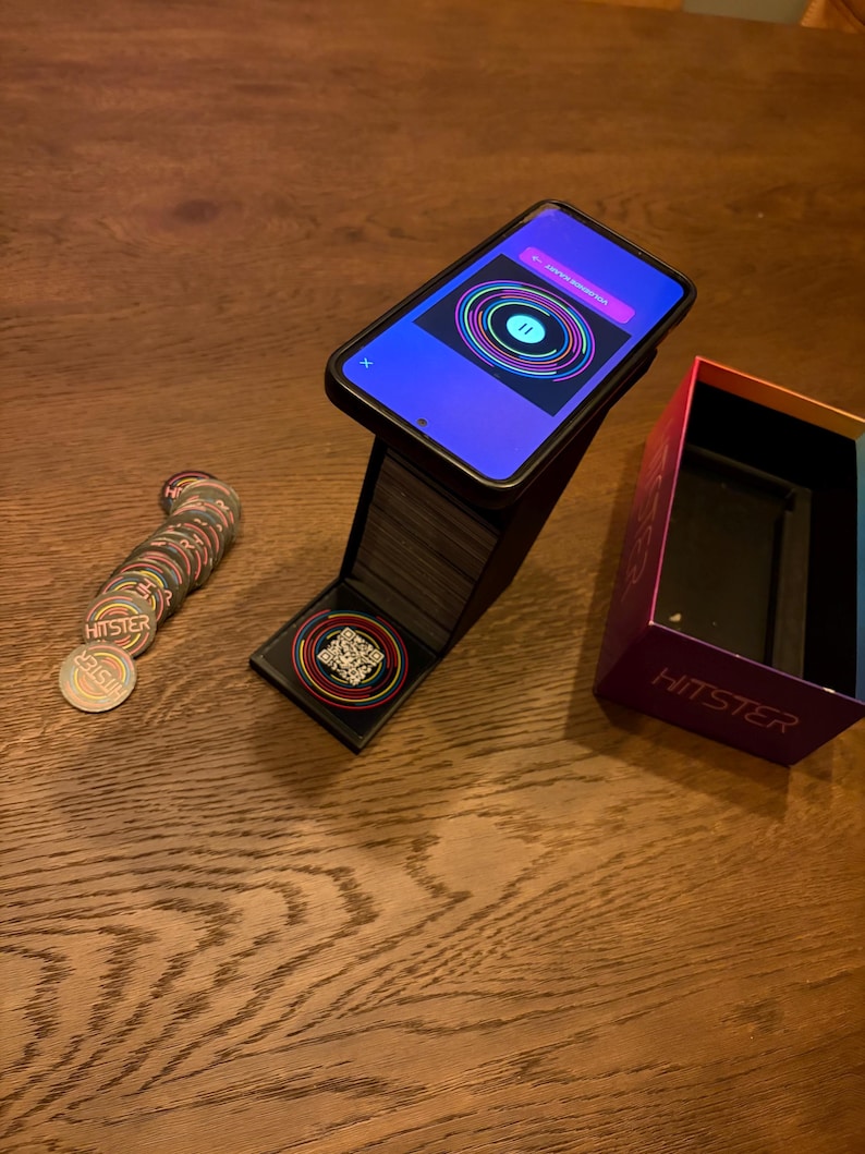 May include: A smartphone showing a colourful circular graphic is propped on a black stand. A stack of circular game pieces with the word "HITSTER" are next to the stand. An open box with the word "HITSTER" is also visible.