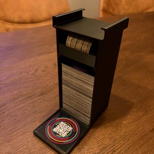 May include: A black card holder containing a stack of cards and a roll of coins. The holder has a hinged lid with a colourful QR code sticker. The holder is on a wooden table.