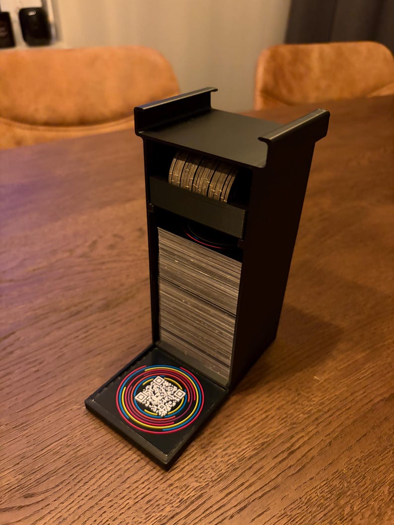 May include: A black card holder containing a stack of cards and a roll of coins. The holder has a hinged lid with a colourful QR code sticker. The holder is on a wooden table.