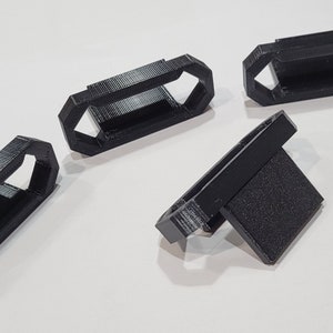 May include: Four black 3D printed plastic parts. Three of the parts are shaped like a rectangle with a hole in the middle. The fourth part is shaped like a rectangle with a small rectangular protrusion on one side.
