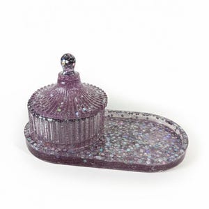 May include: A purple glitter resin trinket box with a matching oval tray. The box has a domed lid with a small, round knob. The tray and box are covered in iridescent glitter.