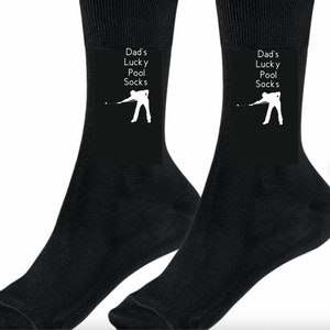 May include: A pair of black socks with a white silhouette of a person playing pool. The socks have the text "Dad's Lucky Pool Socks" on them.