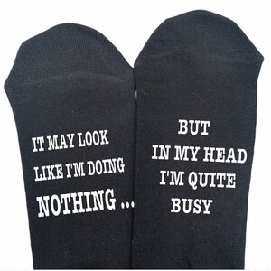 May include: A pair of black socks with white text. One sock reads "IT MAY LOOK LIKE I'M DOING NOTHING..." and the other sock reads "BUT IN MY HEAD I'M QUITE BUSY".