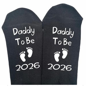 May include: Black socks featuring the text "Daddy To Be" in white, accompanied by white baby footprints and the year "2026". These socks are a gift for a soon-to-be father.