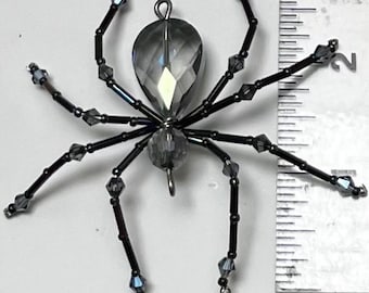 May include: A decorative spider ornament made of faceted gray beads and black wire. The spider has eight legs and a large, teardrop-shaped body. The ornament is approximately 2 inches long.