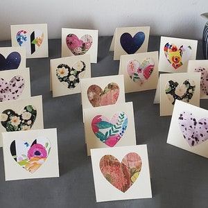 May include: A collection of small, square, cream-colored folded cards, each featuring a heart-shaped cutout on the front. The cutouts reveal various patterns underneath, including floral prints, dark blue starry night designs, and speckled textures.