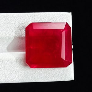 May include: A vibrant red gemstone with a square emerald cut. The gemstone is displayed on a white surface.