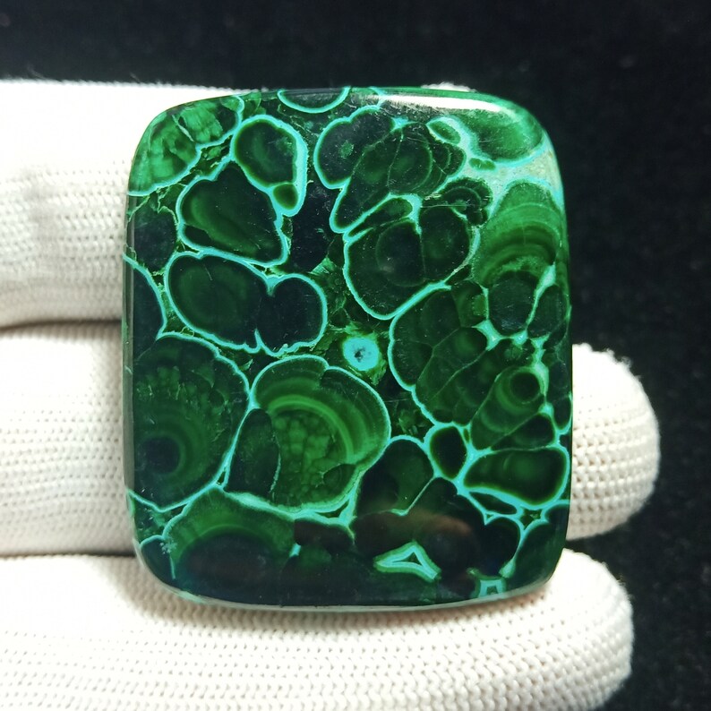 May include: A green malachite cabochon with a swirling, patterned design. The stone has a smooth, polished surface and is a vibrant shade of green.