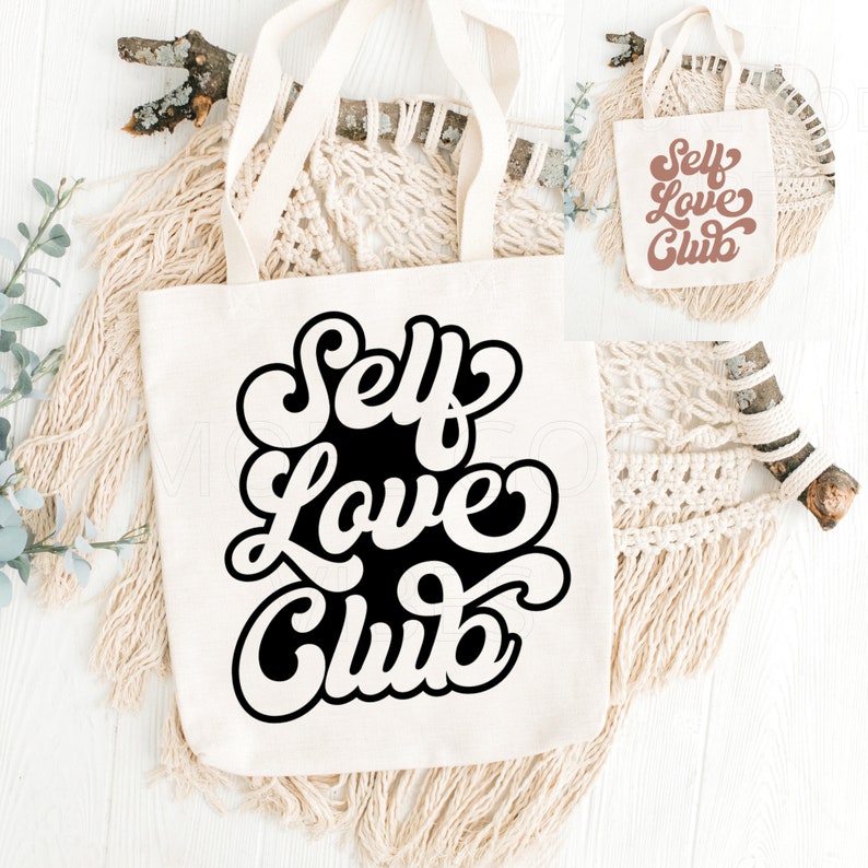 Puede incluir: Bolsa de lona blanca con texto negro que dice "Self Love Club" en un estilo de fuente retro. La bolsa se muestra sobre una superficie de madera blanca con una pared de macram&eacute; y una rama.