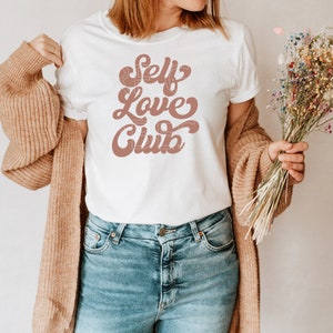 Puede incluir: Camiseta blanca con un gr&aacute;fico marr&oacute;n que dice "Self Love Club" en una fuente retro.