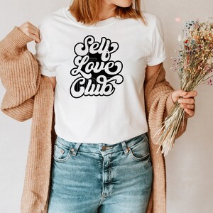 Puede incluir: Camiseta blanca con texto negro que dice "Self Love Club" en un estilo de fuente retro. La camiseta est&aacute; siendo modelada por una persona que lleva jeans azules y una chaqueta de punto marr&oacute;n.