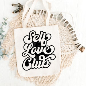Puede incluir: Una bolsa de tela blanca con texto negro que dice "Self Love Club" en un estilo de fuente retro. La bolsa est&aacute; sobre una superficie blanca con una pieza de macram&eacute; tejida y una rama.