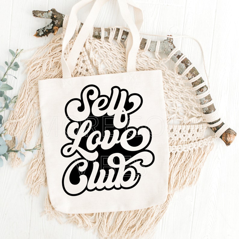 Puede incluir: Una bolsa de tela blanca con texto negro que dice "Self Love Club" en un estilo de fuente retro. La bolsa est&aacute; sobre una superficie blanca con una pieza de macram&eacute; tejida y una rama.