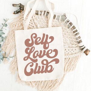 Puede incluir: Una bolsa de lona blanca con asas marrones y el texto "Self Love Club" en una fuente marr&oacute;n de estilo retro.