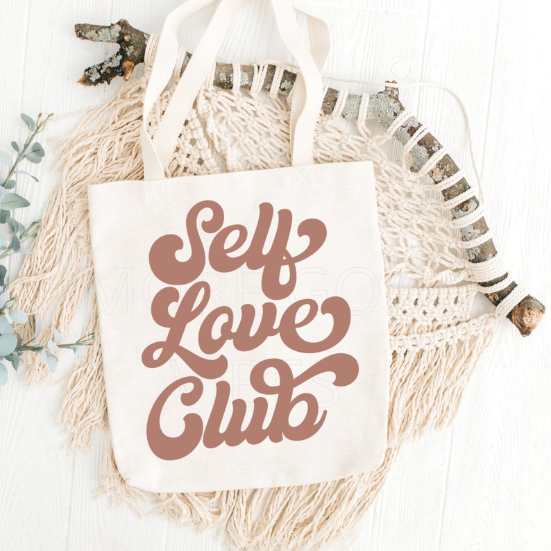 Puede incluir: Una bolsa de lona blanca con asas marrones y el texto "Self Love Club" en una fuente marr&oacute;n de estilo retro.