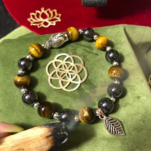 May include: A beaded bracelet with tiger's eye and hematite beads, a silver Buddha charm, and a silver leaf charm. The bracelet is on a green velvet surface.