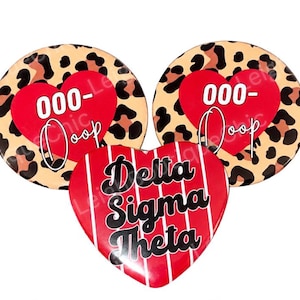 May include: Two circular buttons featuring a tan and black leopard print background with a red heart and the text 000-Ooop, and one heart-shaped button with a red background, white vertical stripes, and the text Delta Sigma Theta in black.