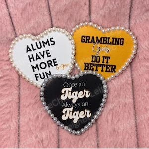 May include: Three heart-shaped plaques bordered with white pearls. The white plaque reads ALUMS HAVE MORE FUN Go Tigers!. The yellow plaque reads GRAMBLING Girls DO IT BETTER. The black plaque reads Once an Tiger Always an Tiger in white text.