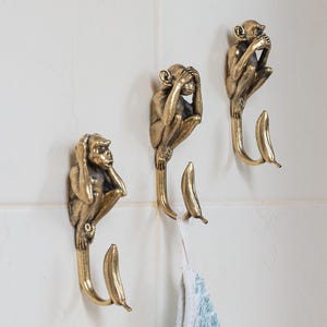 May include: Three brass-colored monkey-shaped wall hooks in a row, each depicting a different pose from the "see no evil, hear no evil, speak no evil" proverb. The hooks are mounted on a white tiled wall, with a blue and white towel hanging from one.