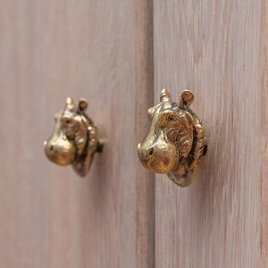 Op de afbeelding: Twee gouden hippopotamusvormige kastdeurknopjes op een lichte houten kastdeur.