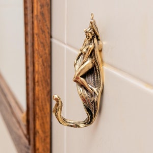 May include: A gold-toned metal hook shaped like a woman with a decorative design. The hook is mounted on a white tiled wall.