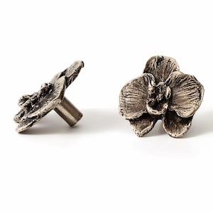 May include: Two metal cabinet knobs shaped like orchid flowers with a dark, weathered silver finish. One knob is shown from a front-facing perspective, while the other is angled to reveal the cylindrical mounting post on the back side.