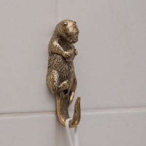 Op de afbeelding: Een messingkleurige wandhaak in de vorm van een staande bever. De haak is bevestigd aan een witte tegelwand en houdt een blauw-wit patroonhanddoek vast. De beverhaak is een decoratieve toevoeging.