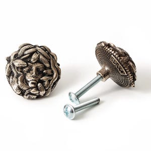 May include: Two round cabinet knobs with an antique silver finish, featuring a detailed relief of a face surrounded by snakes. Each knob has a threaded metal post and comes with a separate silver-colored screw for installation on furniture.