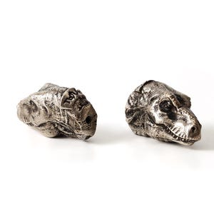 May include: Two metallic silver-toned beads shaped like dinosaur skulls. The bead on the left depicts a triceratops with three horns, while the bead on the right depicts a tyrannosaurus rex with a detailed jaw, teeth, and eye sockets.