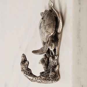 May include: A silver-toned metal wall hook featuring a detailed sea turtle design. The turtle is positioned above a textured, coral-like hook structure. The metallic finish has a weathered appearance with visible shell patterns and flippers.