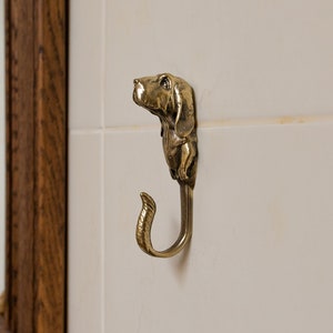 May include: A gold-toned metal hook shaped like a dog's head. The hook is mounted on a white tiled wall.