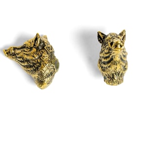 May include: Two small, detailed brass-colored boar head figurines. One is angled to the left, showing the side profile, and the other faces forward. The figurines are on a white background.