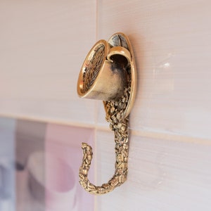 May include: A gold-toned metal hook shaped like a coffee cup with a decorative design. The hook is mounted on a white tiled wall.
