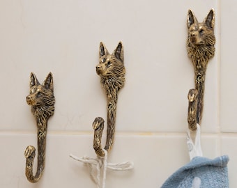 May include: Three gold-toned fox head hooks mounted on a white tiled wall. A blue towel hangs from one of the hooks.
