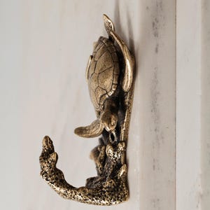 Op de afbeelding: Een wandhaak van antiek messingkleurig metaal met een gedetailleerd ontwerp van een zeeschildpad. De schildpad bevindt zich boven een gestructureerde, koraalachtige haakvorm die naar boven buigt en dient als functioneel ophangpunt.