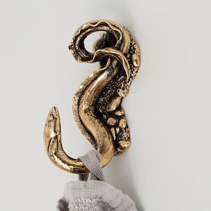 Puede incluir: Un gancho de pared de metal dorado, con forma de anguila enroscada y una superficie texturizada que recuerda a rocas. Una toalla de algodón de color gris claro y textura suave cuelga de la parte inferior curva del gancho.