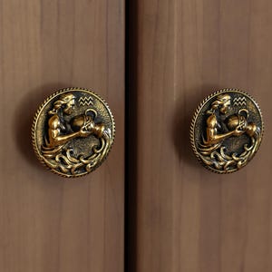 Puede incluir: Dos tiradores de armario de latón antiguo con un diseño en relieve. Cada tirador tiene forma circular con un relieve detallado de una figura sosteniendo un recipiente, sobre un fondo texturizado. Los tiradores están montados en un armario de madera marrón claro.