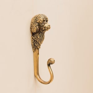 May include: A gold-toned metal hook shaped like a poodle's head. The hook is mounted on a white wall.