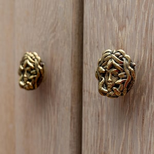 Peut inclure: Deux boutons de porte de placard dorés avec un design détaillé d'un visage de femme entouré de serpents.