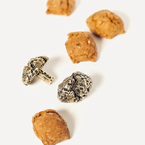 Peut inclure: Un ensemble de six petits biscuits dorés, certains avec une texture rugueuse, et deux objets en métal dorés. L'un des objets en métal est en forme de champignon avec le mot "MAUKA" inscrit sur le pied.
