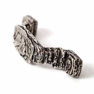 May include: A silver-toned metal cabinet pull with a highly textured, rugged surface that mimics the appearance of natural tree bark. The handle features an irregular, organic shape with deep grooves and raised ridges throughout its length.